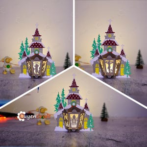 Könnte beinhalten: Eine Weihnachtskirche aus Papier mit einem Licht im Inneren. Die Kirche ist mit einem Kranz und Schnee verziert. Auf beiden Seiten der Kirche stehen Bäume.