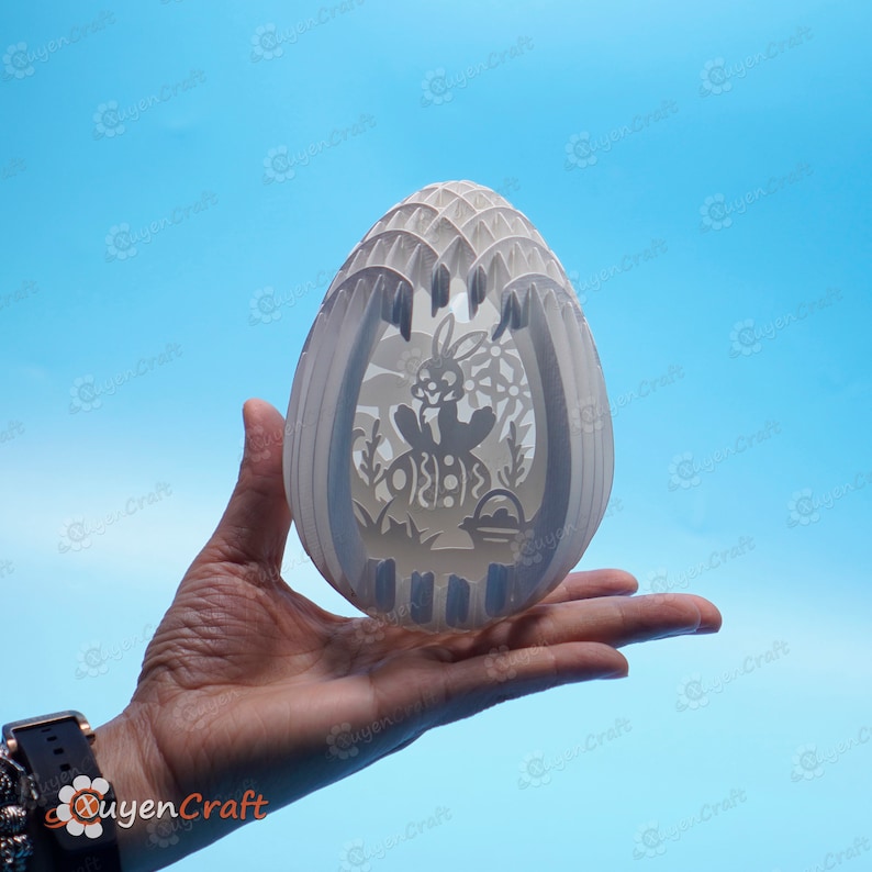 May include: A white papercraft Easter egg with a cut-out design of a bunny holding a basket. The egg is held in a person's hand.