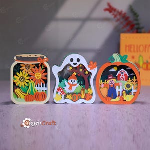 Könnte beinhalten: Drei geschichtete Holzdekorationen mit Herbstmotiven: ein Einmachglas mit Sonnenblumen und Kürbissen, ein Geist im Wald mit Pilzen und Herbstblättern sowie ein Kürbisrahmen mit einer Vogelscheuche, einer roten Scheune und Sonnenblumen.