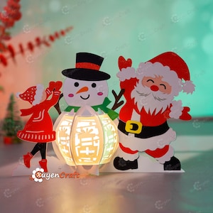 May include: A white snowman wearing a black top hat, a red and white girl holding a red heart, and a red and white Santa Claus with a white beard. The figures are standing on a white base with a white lantern in the centre. The lantern has a white light shining through the cutouts.