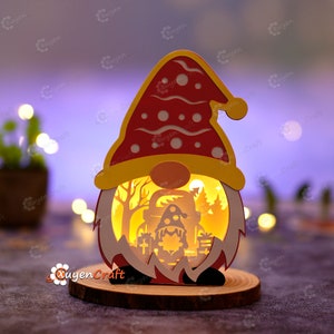 Pode incluir: Uma luz decorativa em forma de gnomo de Natal. O gnomo tem um chap&eacute;u vermelho com bolinhas brancas, uma aba amarela e um nariz redondo laranja. A cena interna retrata uma floresta com um gnomo menor, &aacute;rvores e presentes. A luz est&aacute; em uma base de madeira.