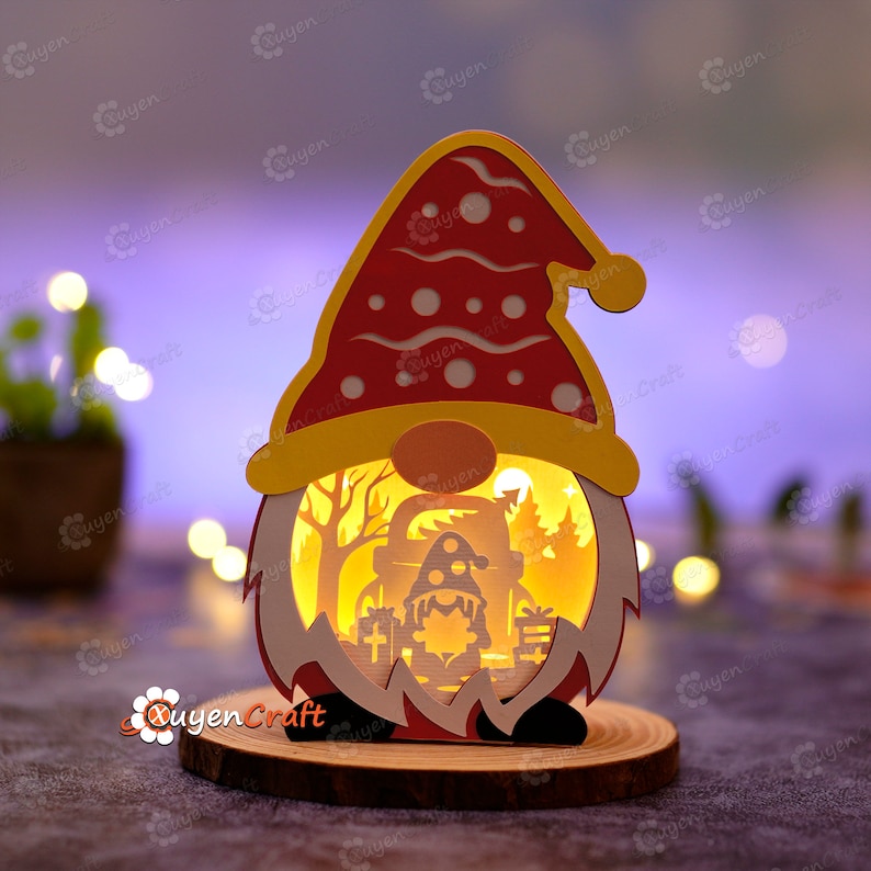Pode incluir: Uma luz decorativa em forma de gnomo de Natal. O gnomo tem um chap&eacute;u vermelho com bolinhas brancas, uma aba amarela e um nariz redondo laranja. A cena interna retrata uma floresta com um gnomo menor, &aacute;rvores e presentes. A luz est&aacute; em uma base de madeira.