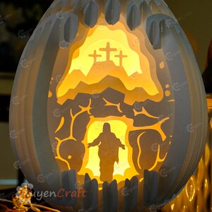 May include: A white paper Easter egg with a 3D paper cut-out design of a figure emerging from a cave with a cross on a hill behind them. The egg is illuminated from within, creating a warm glow.