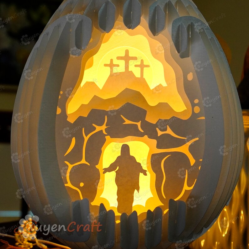 May include: A white paper Easter egg with a 3D paper cut-out design of a figure emerging from a cave with a cross on a hill behind them. The egg is illuminated from within, creating a warm glow.
