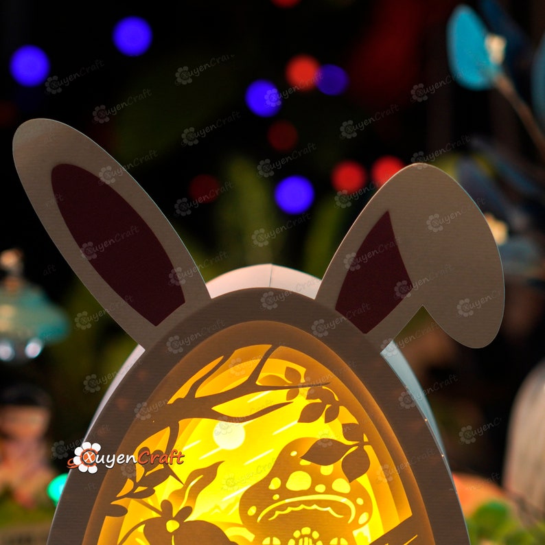 May include: A white papercraft Easter egg shaped nightlight with bunny ears. The egg has a cut-out design of a forest scene with a mushroom and a moon. The light is on and illuminates the scene.