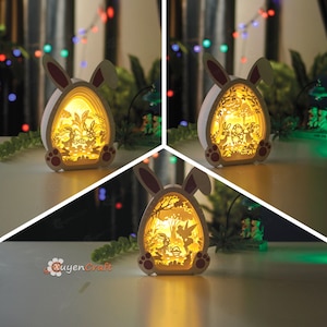 Könnte beinhalten: Eine weiße, hölzerne Osterhasen-Nachtlampe mit einem dreidimensionalen, mehrschichtigen Design. Der Hase hält ein Ei und im Ei befinden sich Blumen und ein Baum. Das Licht wird mit einem warmen gelben Schein beleuchtet.