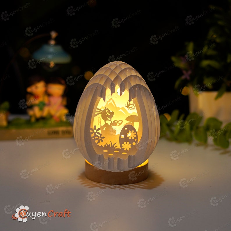 May include: A white, paper-crafted Easter egg night light with a cut-out design of a bunny rabbit, flowers, and an egg. The light is illuminated from within, casting a warm glow.