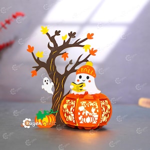 May include: A Halloween papercraft decoration. It features a tree with autumn leaves, a ghost, and an orange pumpkin lantern. The lantern has a ghost wearing a hat with "FALL" on it, reading a book. A small pumpkin is also present.