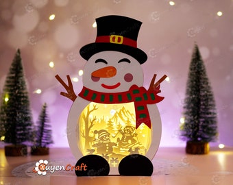 May include: A white snowman with a red and green scarf and a black top hat. The snowman has a light inside that illuminates a winter scene with two smaller snowmen.