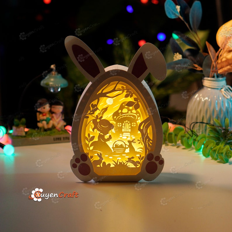 May include: A white wooden Easter egg shaped night light with a bunny rabbit design. The light features a cut-out scene of a rabbit and a girl in a forest with a mushroom house. The light is illuminated with a warm yellow glow.