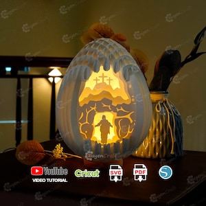 May include: A white papercraft Easter egg lamp with a 3D design of Jesus emerging from a tomb. The egg is illuminated from within, casting a warm glow. The lamp is on a wooden base and there are icons for YouTube, Cricut, SVG, PDF, and a silhouette of a person.