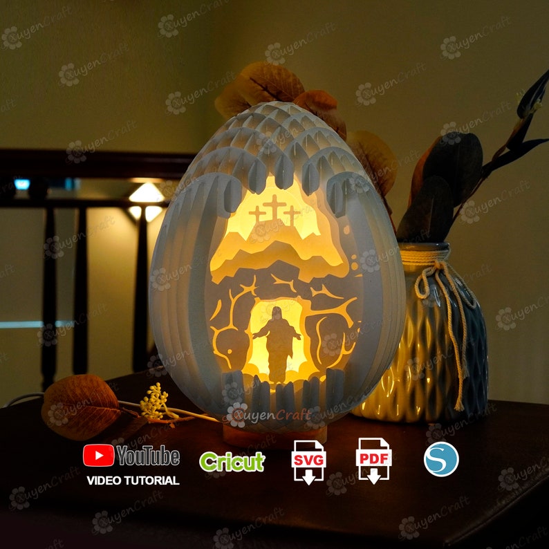 May include: A white papercraft Easter egg lamp with a 3D design of Jesus emerging from a tomb. The egg is illuminated from within, casting a warm glow. The lamp is on a wooden base and there are icons for YouTube, Cricut, SVG, PDF, and a silhouette of a person.