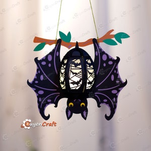 May include: A black and purple paper bat lantern with a white light inside. The bat is hanging from a branch with green leaves.