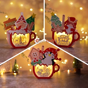 Peut inclure: Trois mugs lumineux sur le thème de Noël avec des personnages différents. Le premier mug représente un Père Noël avec une pancarte qui dit "Xmas". Le deuxième mug représente un gnome avec une pancarte qui dit "Xmas". Le troisième mug représente un pingouin avec une canne en sucre.