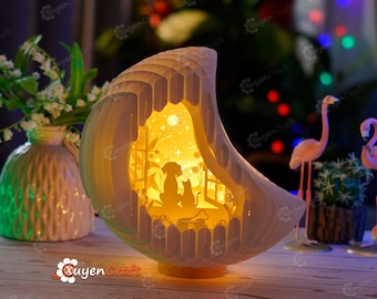 May include: A white papercraft lamp shaped like a crescent moon with a cut-out design of a dog and cat sitting by a window. The lamp is lit from within, casting a warm glow. The lamp is on a wooden table with a vase of white flowers and a potted plant with pink flamingos in the background.