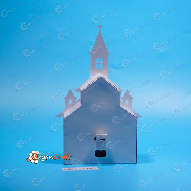 May include: White paper model of a church with a cross on top. The model has a small window in the front and a door on the side. The model is sitting on a blue surface.