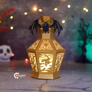 May include: A gold-colored paper lantern with a black bat on top. The lantern has cut-out designs of spiders, a witch, and a haunted house. The lantern is lit from within, creating a warm glow. The lantern is a Halloween decoration.