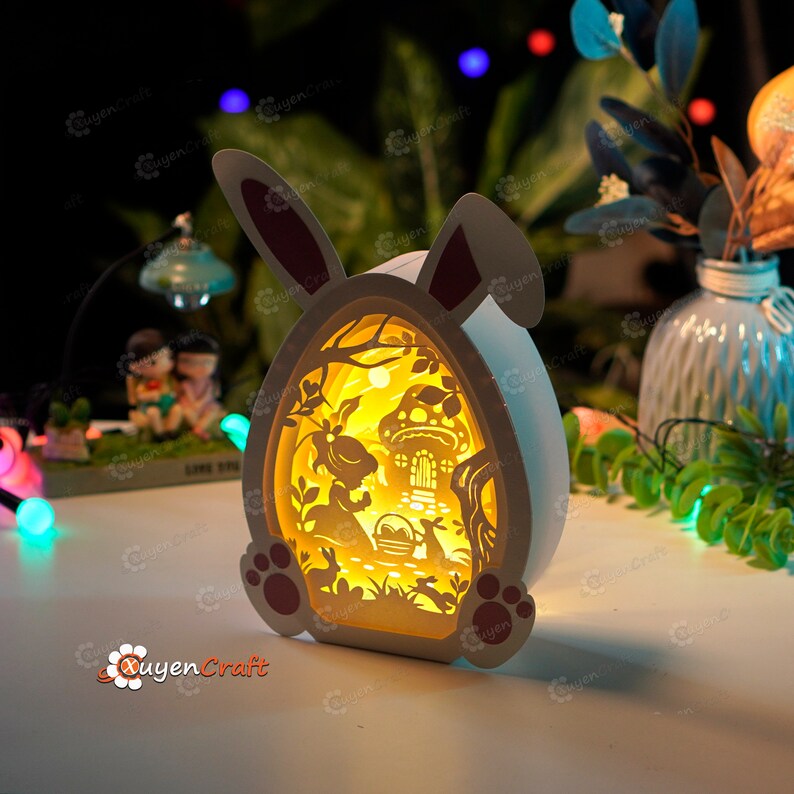 May include: A white wooden Easter bunny night light with a cut-out design of a bunny in a forest scene. The light is illuminated from the inside, creating a warm glow.