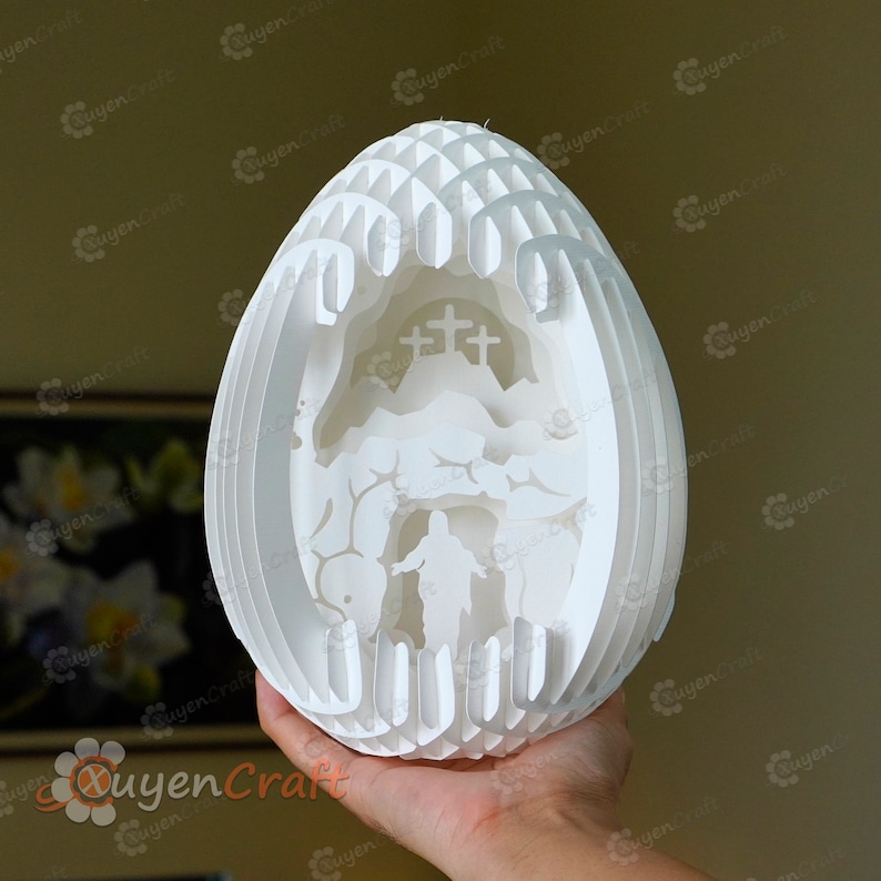 May include: A white papercraft Easter egg with a 3D design depicting the resurrection of Jesus. The egg is made of multiple layers of paper that are cut and folded to create a detailed and intricate design. The design includes a cross, a hill, and a figure of Jesus walking out of a tomb.