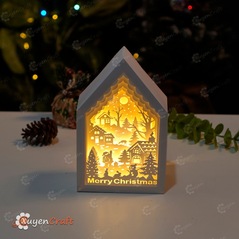 May include: White wooden house shaped Christmas light with a cut-out design of a winter village scene. The light illuminates the scene with a warm glow. The words "Merry Christmas" are printed on the bottom of the house.