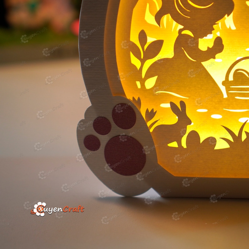 May include: A white papercraft lamp with a cut-out design of a girl holding a basket of eggs, a rabbit, and flowers. The lamp is lit from within, casting a warm glow. The lamp has a paw print design on the bottom.