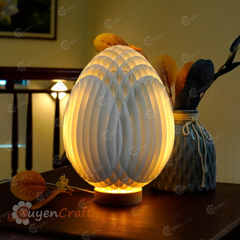 May include: A white egg-shaped lamp with a geometric pattern made of overlapping layers. The lamp is lit and casts a warm glow. It is sitting on a wooden table next to a gray vase with dried leaves.