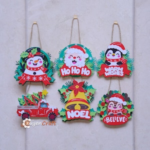 Op de afbeelding: Zes decoratieve hangende ornamenten met kerstthema's, waaronder een sneeuwman, de Kerstman met de tekst Ho Ho Ho, een pinguïn met Warm Wishes, een rode vrachtwagen, een gele bel met Noel en een rendier met het woord Believe aan een touwtje.