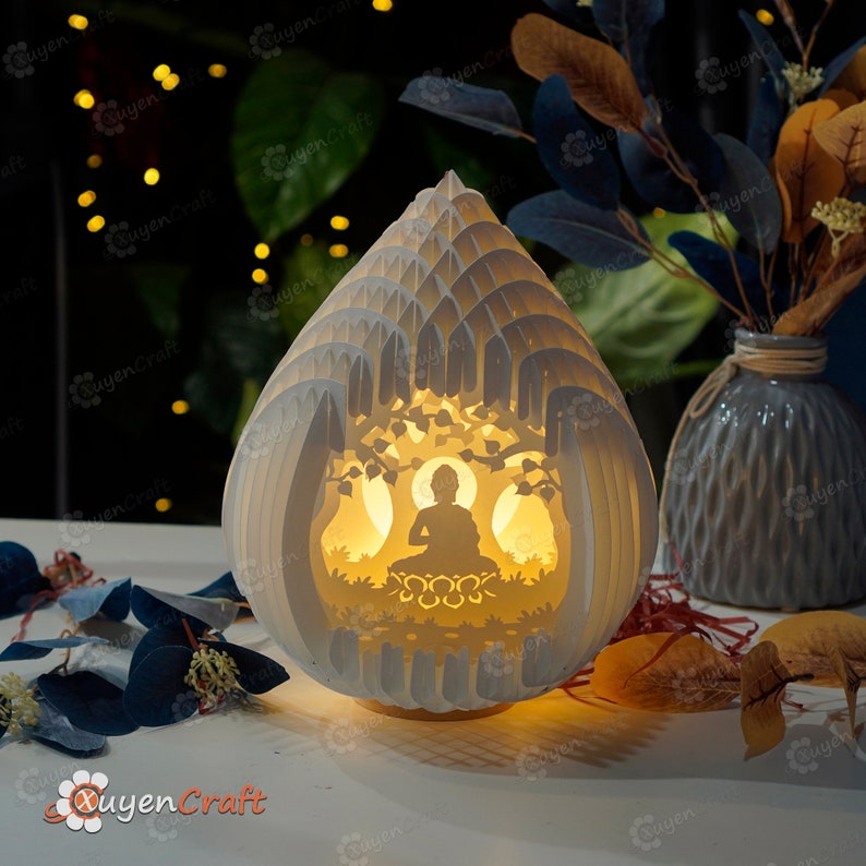 Op de afbeelding: Een witte lamp van papierkunst met een silhouet van Boeddha die onder een boom mediteert. De lamp wordt van binnenuit verlicht, waardoor een warm gloed ontstaat.