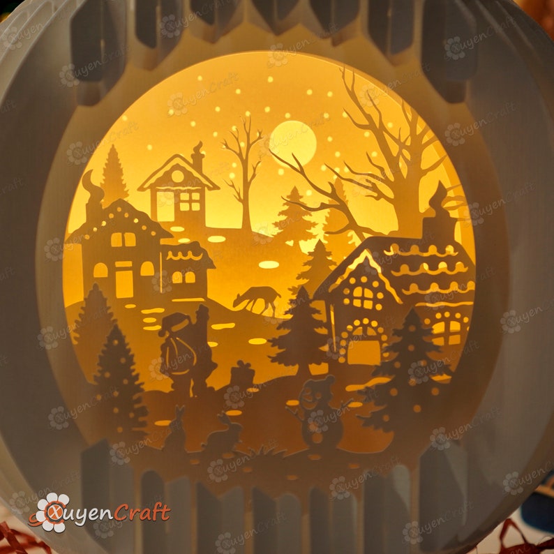 May include: A white papercraft light-up ornament with a winter scene. The scene includes a small village with houses, trees, a deer, and Santa Claus. The ornament is illuminated from behind, creating a warm glow.