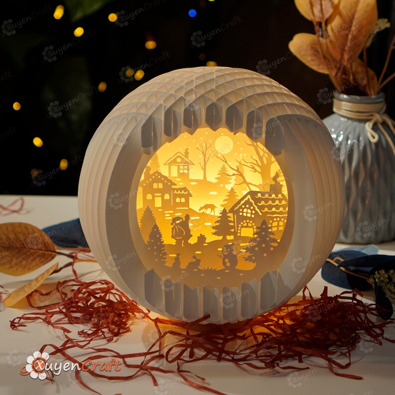 May include: A white papercraft sphere with a cut-out design of a winter village scene. The scene includes houses, trees, a snowman, and a deer. The sphere is illuminated from within, creating a warm glow.
