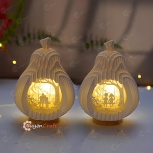 May include: Two pear-shaped, white paper lamps with a warm glow. Each lamp features a cut-out scene of a couple on a swing under a tree. The lamps sit on a light-colored base. The brand name "Quyen Craft" is visible.