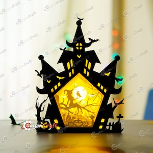 May include: A black papercraft Halloween haunted house with a witch and a crow on a branch, a full moon, and a graveyard scene inside. The house is illuminated from behind, creating a glowing effect.