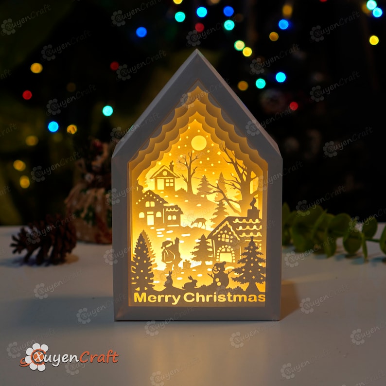 May include: A white wooden Christmas light-up house with a cut-out design of a winter village scene. The house has a warm yellow glow and the words "Merry Christmas" are printed on the front.