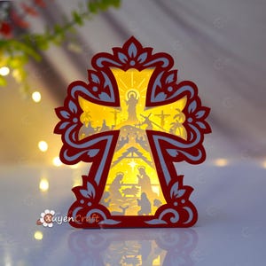 May include: A layered paper craft cross featuring a red and white ornate frame with a backlit yellow center. The design depicts a nativity scene at the bottom and a crucifixion scene at the top, with the text XuyenCraft visible in the lower left corner.