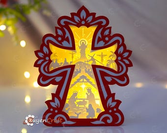 May include: A layered paper craft cross featuring a red and white ornate frame with a backlit yellow center. The design depicts a nativity scene at the bottom and a crucifixion scene at the top, with the text XuyenCraft visible in the lower left corner.