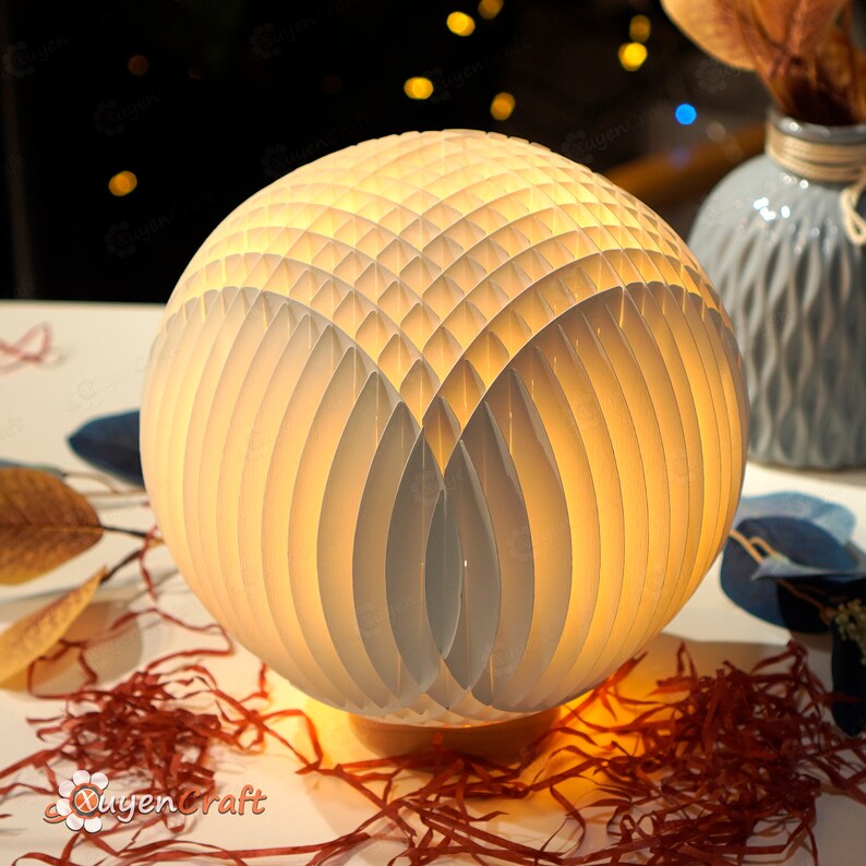 May include: A white paper lamp with a geometric design. The lamp is lit and casting a warm glow. The lamp is sitting on a table with brown paper shreds.