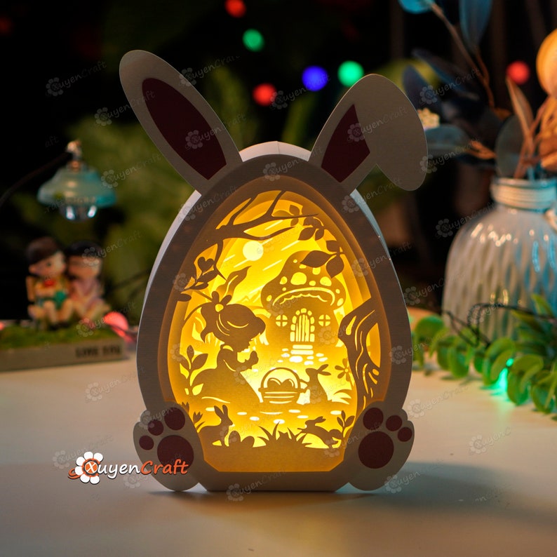 May include: A white bunny-shaped Easter egg night light with a cut-out design of a girl and a bunny in a forest setting. The light is illuminated from within, casting a warm glow.