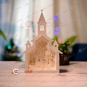 May include: A white papercraft model of a church with a snowy forest scene inside. The church has a steeple with a cross on top and windows. The forest scene includes trees, a deer, and a small fence.