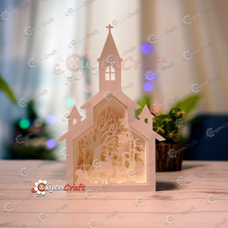 May include: A white papercraft model of a church with a snowy forest scene inside. The church has a steeple with a cross on top and windows. The forest scene includes trees, a deer, and a small fence.
