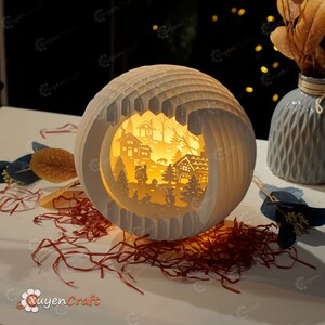 May include: A white papercraft sphere with a cut-out design of a winter village scene. The sphere is illuminated from within, creating a warm glow. The scene includes houses, trees, and a figure of Santa Claus.