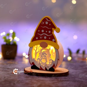 Pode incluir: Uma lanterna de papel com tema natalino em forma de gnomo, com chap&eacute;u e barba vermelhos e detalhes amarelos. O interior &eacute; iluminado, revelando uma cena festiva. Repousa sobre uma base de madeira.