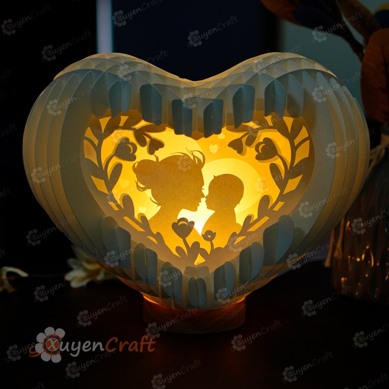 May include: A white paper heart-shaped lamp with a cutout design of a mother and child. The lamp is illuminated from within, casting a warm glow on the silhouettes. The design features delicate floral details and a moon.