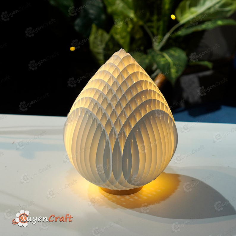 Op de afbeelding: Een witte, geometrische tafellamp met een uniek, gelaagd ontwerp dat lijkt op een bloemknop. De lamp is verlicht en geeft een warm licht.