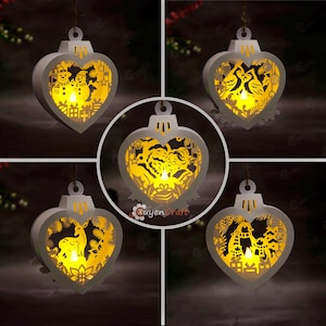May include: Five heart-shaped white wooden ornaments with intricate laser-cut designs illuminated by warm yellow internal lights. The cutouts feature holiday themes including snowmen, birds, Santa Claus, a deer, and penguins with gifts.