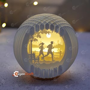 May include: A spherical white paper-cut light box featuring a layered silhouette scene of two people jogging in a park. The interior is illuminated with a warm yellow glow, highlighting a bench, a lamp post, trees, and a distant city skyline.