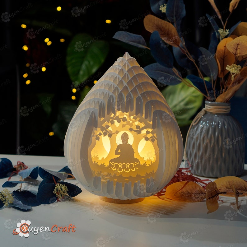 Op de afbeelding: Een witte lamp van papierkunst in de vorm van een traan met een uitgesneden ontwerp van een mediterend Boeddha-figuur in een boom. De lamp is verlicht en geeft een warm licht.