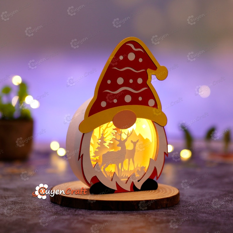 Pode incluir: Uma lanterna em forma de gnomo natalino, com chap&eacute;u vermelho e barba branca. A lanterna apresenta uma cena de floresta com veados e &aacute;rvores iluminadas por dentro. Repousa sobre uma base de madeira, criando um brilho acolhedor.