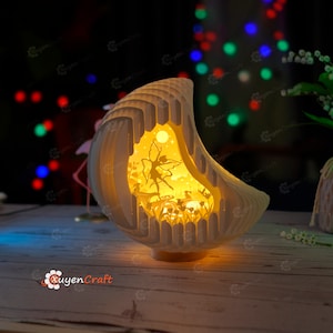 May include: A white, crescent moon-shaped lamp with a fairy and flowers cut out design. The lamp is lit and casts a warm glow. The lamp is on a wooden table.