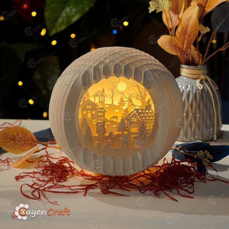 May include: A white papercraft sphere with a cut-out design of a winter village scene. The scene includes houses, trees, a moon, and a couple walking through the snow. The sphere is illuminated from within, creating a warm glow.