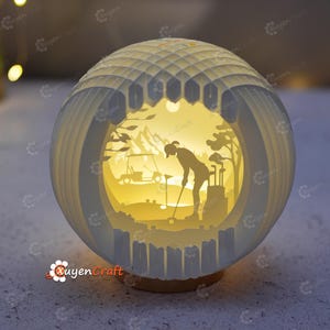 Może przedstawiać: Sferyczna lampa z papieru przedstawiająca scenę z golfa. Lampa ma warstwową konstrukcję z sylwetkami golfisty, wózka golfowego i drzew na tle ciepłego, świecącego tła. Zewnętrzna powłoka jest biała z geometrycznym wzorem i stoi na drewnianej podstawie.